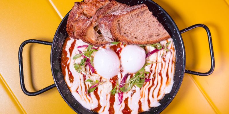 Shakshuka con guiso de pimiento rojo, tomate, harissa, dos huevos poché ecológicos, salsa de yogur con comino, queso feta, cebolla encurtida y tostadas de pan de Pagés de masa madre en Berry Brunch Barcelona.